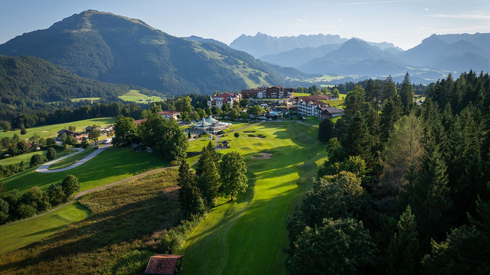 Luftaufnahme: Ein Fairway zieht sich durch den Wald auf ein Resort zu, dahinter Berge im Abendlicht