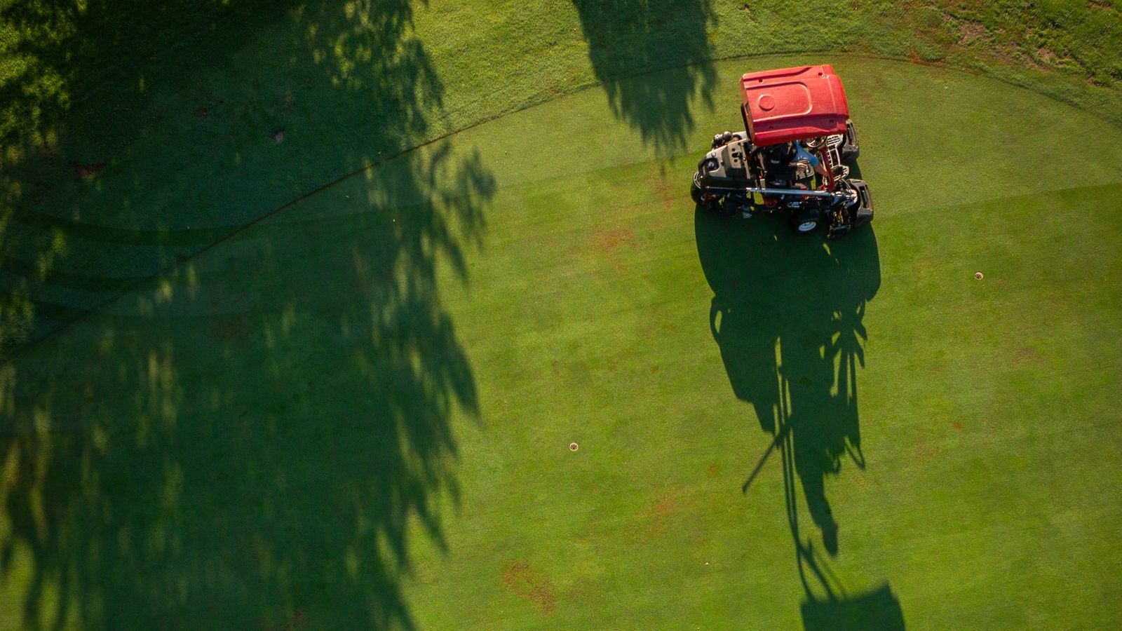 Luftaufnahme von oben: Ein Greenkeeper mäht frühmorgens ein Grün, lange Baumschatten liegen über dem Rasen